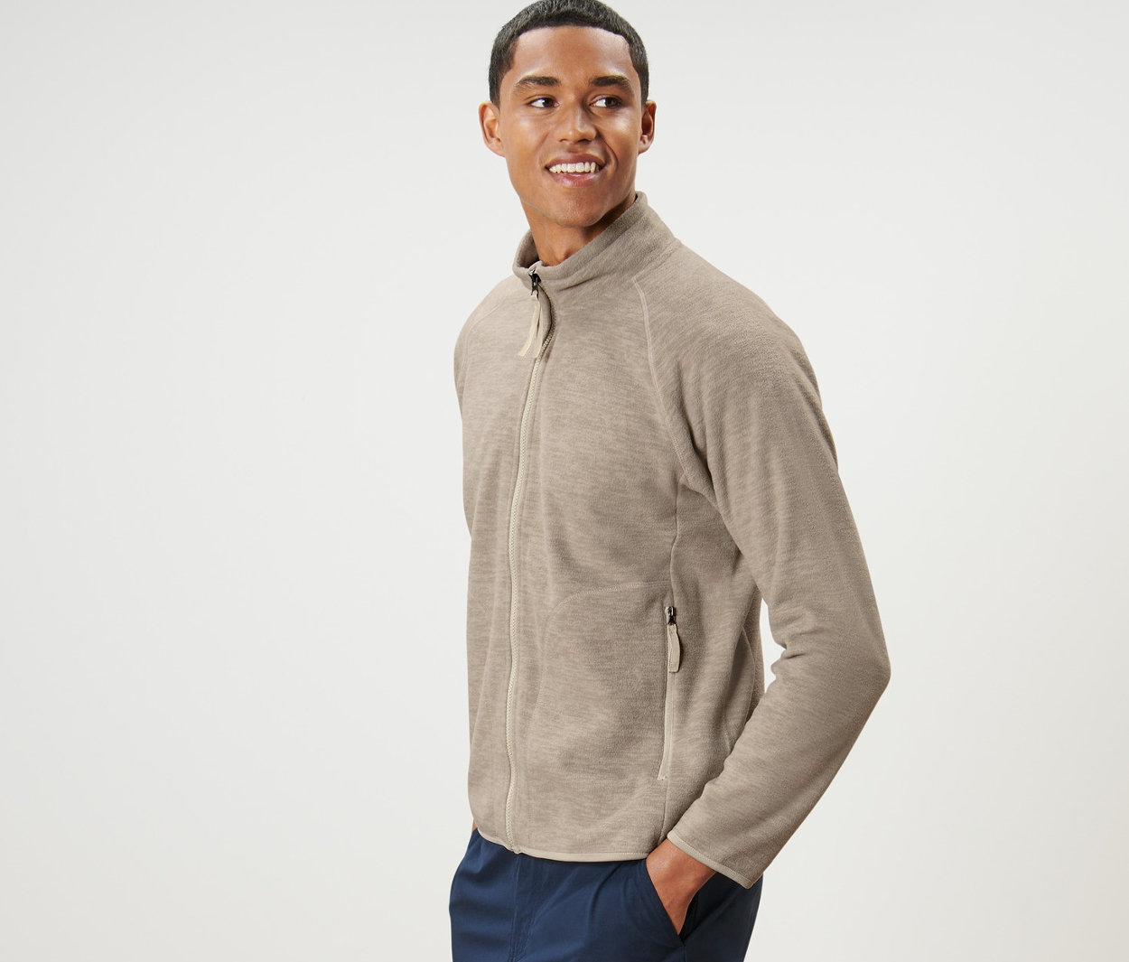 Muž pózuje v béžovej flísovej bundě a tmavomodrých nohaviciach.