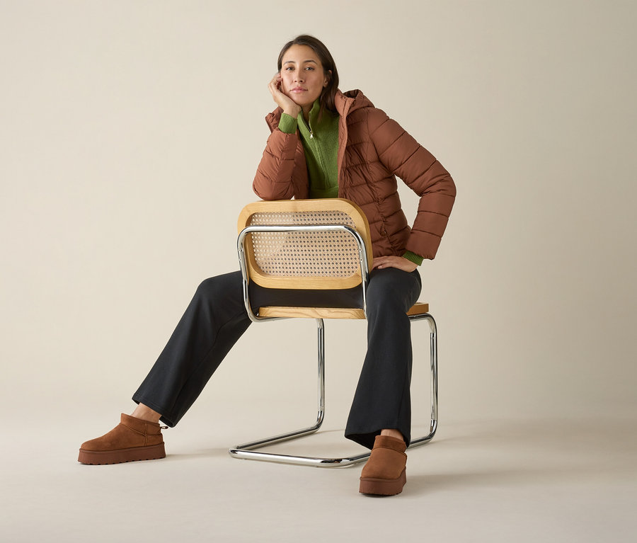 Žena sedí na stoličke v pletenom svetri Troyer, bunde a topánkach.