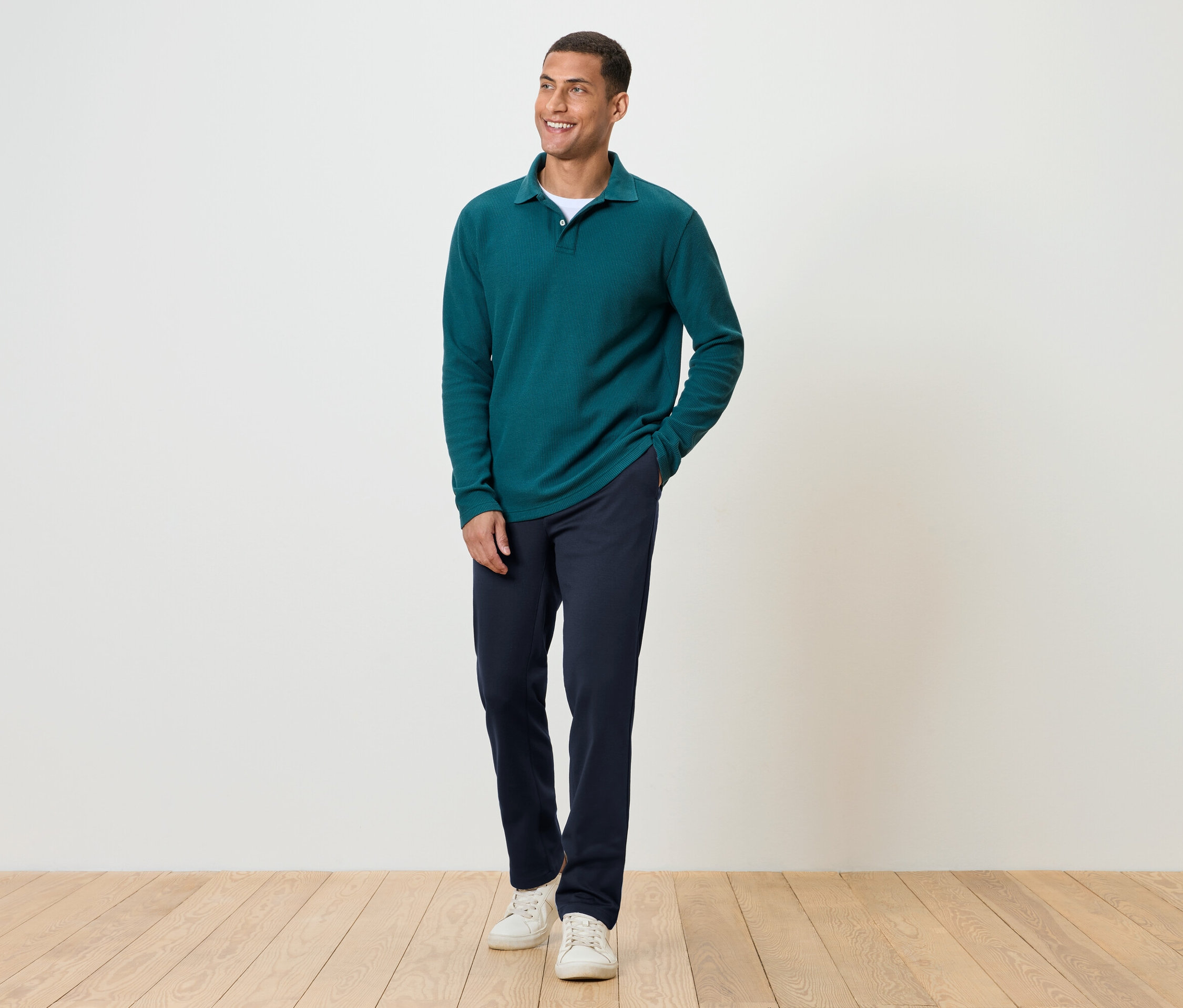 Muž stojí na drevenej podlahe a má na sebe tmavomodré nohavice, zelené tričko s dlhým rukávom a biele tenisky.