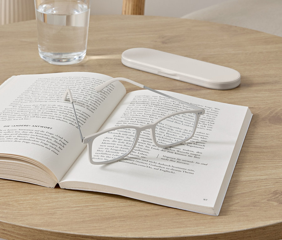 Okuliare ležia na otvorenej knihe na okrúhlom stolíku. Vedľa knihy stojí pohár s vodou a puzdro na okuliare.