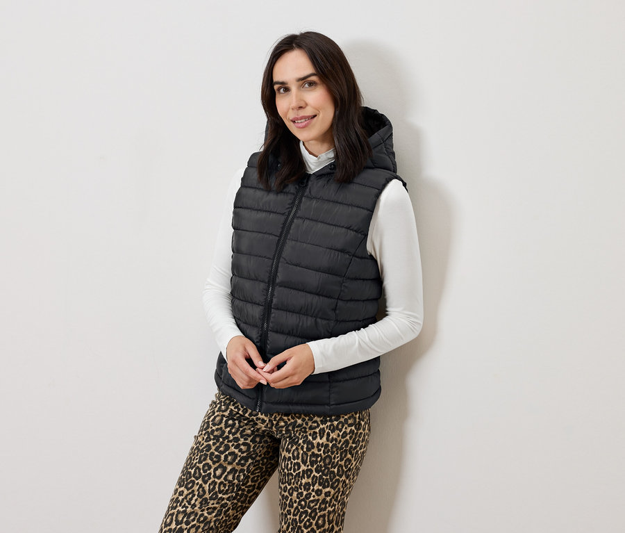 Žena pózuje v čiernej prešívanej veste s kapucňou, bielom roláku a zvieracích nohaviciach.