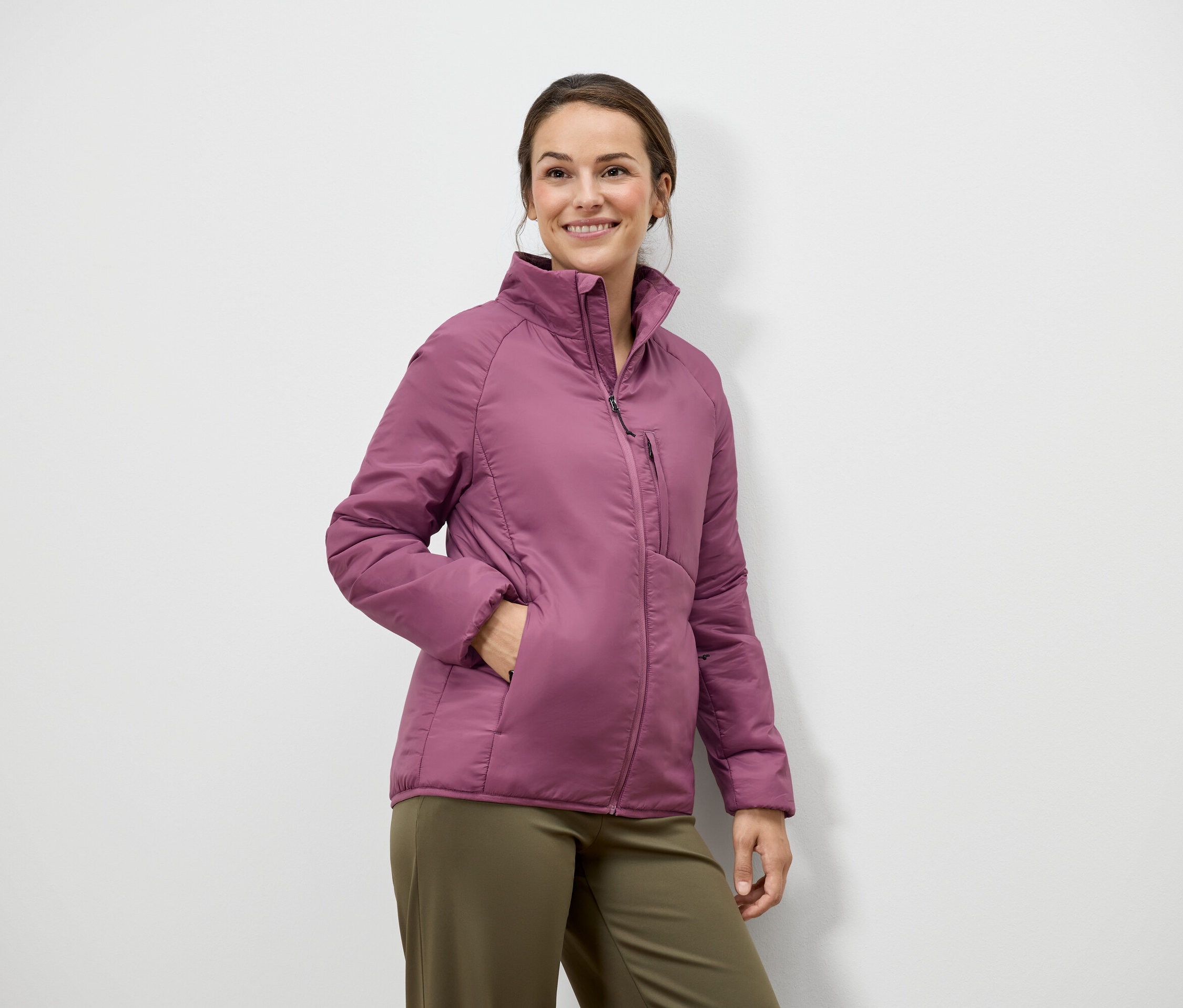 Žena s hnedými vlasmi a úsmevom pózuje v ružovej bunde a zelených nohaviciach pred bielou stenou.