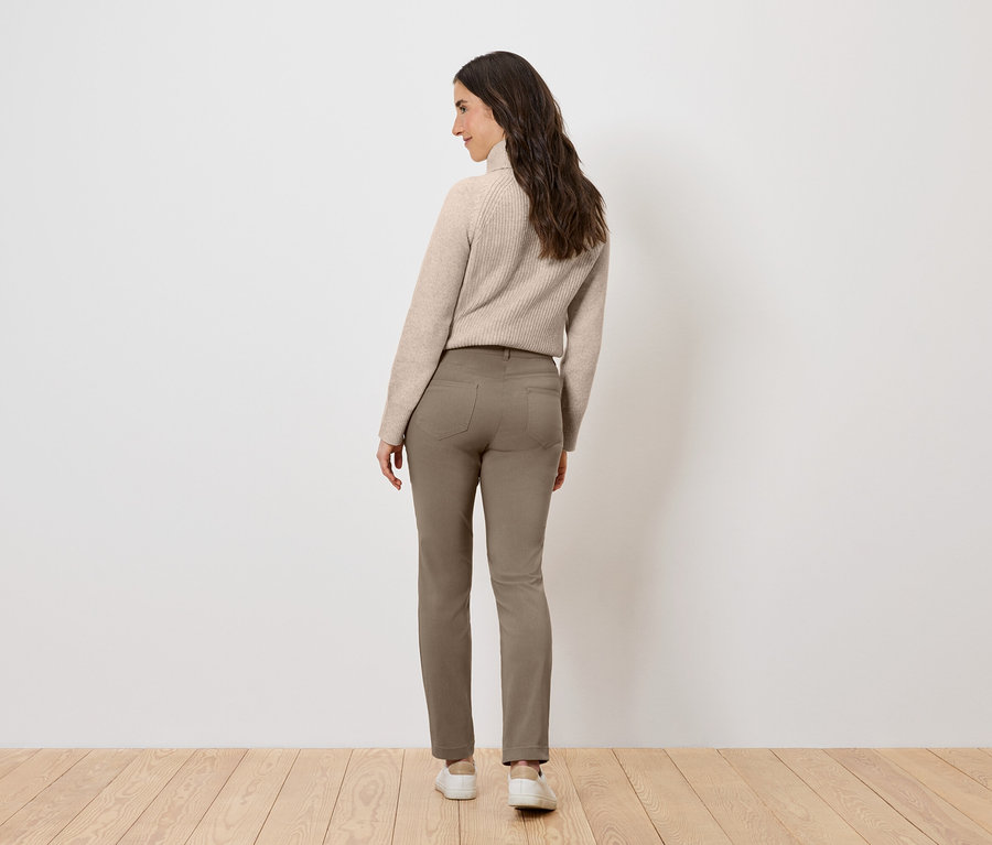 Žena v béžovom svetri a hnedých nohaviciach, stojaca chrbtom k divákovi.