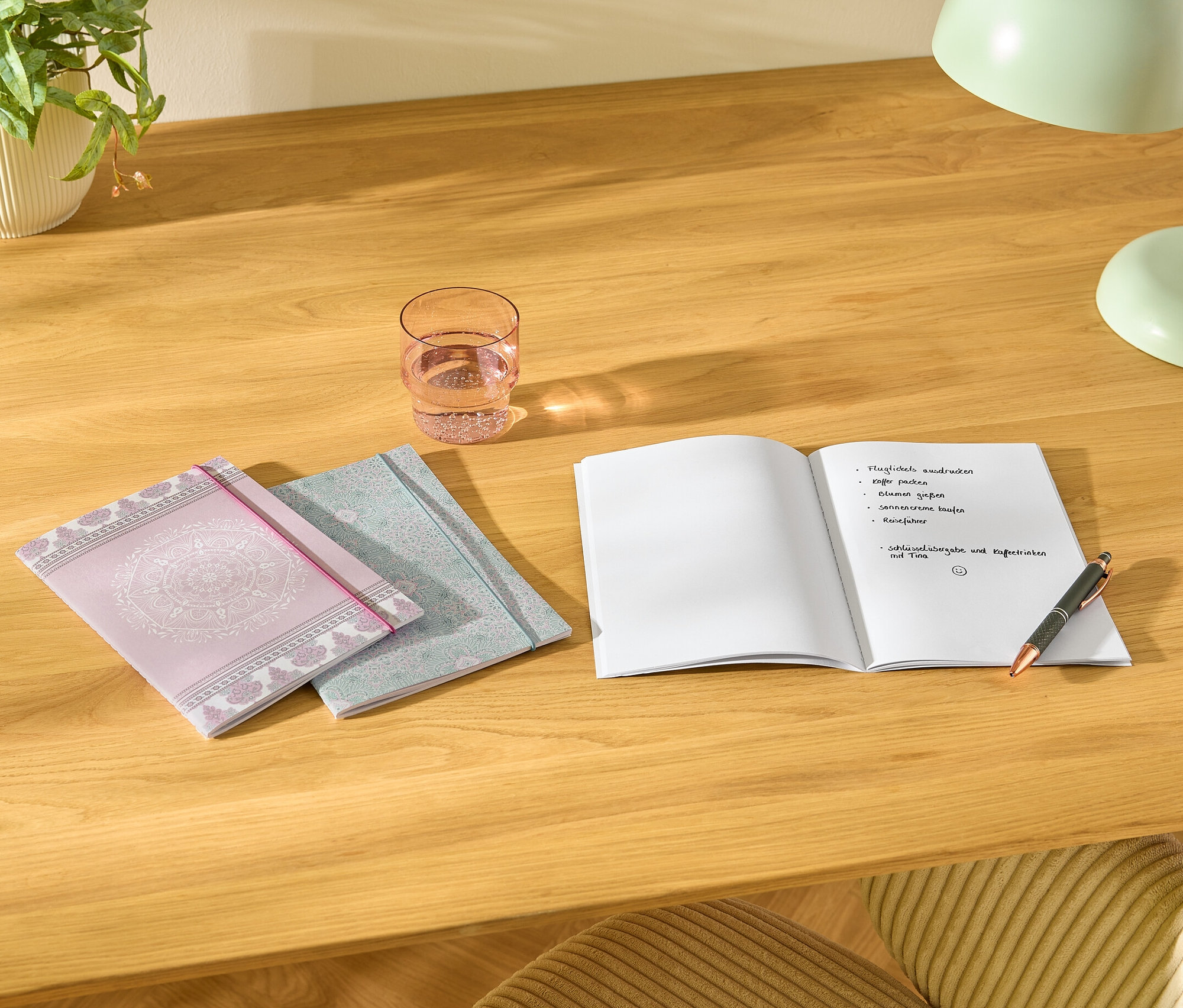 Na stole ležia tri Notizbücher, pohár vody, otvorený zápisník s poznámkami a pero.