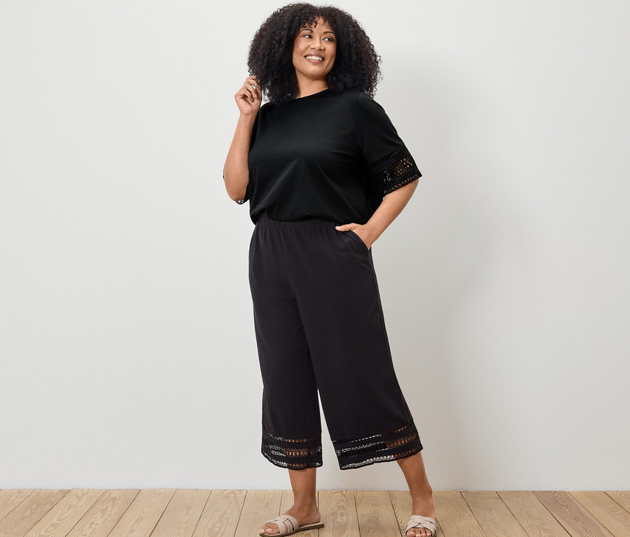 Žena pózuje v čiernom tričku s výšivkou a čiernych nohaviciach culottes s výšivkou.