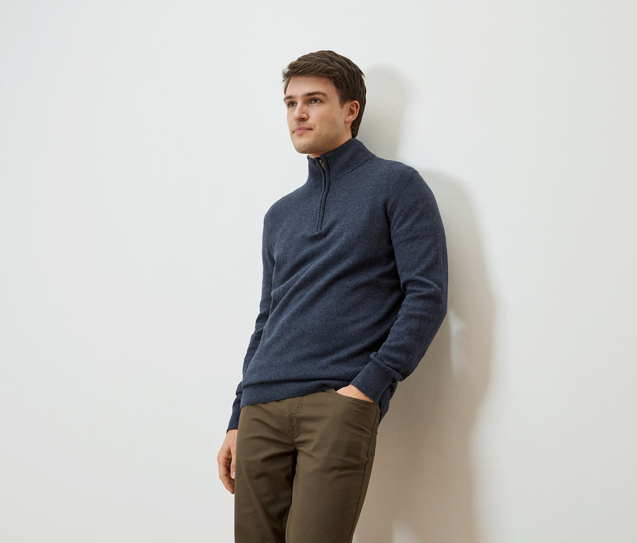 Muž sa opiera o stenu, má na sebe tmavomodrý pulóver na zips a khaki nohavice.