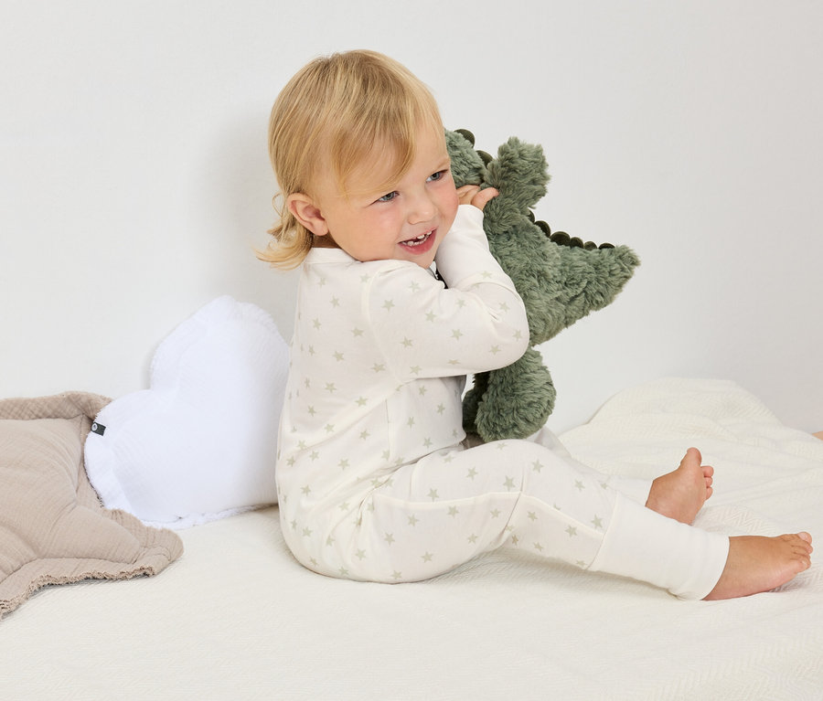 Blondínka v bielom pyžame s hviezdami sedí na bielej deke a objíma zeleného plyšového dinosaura.