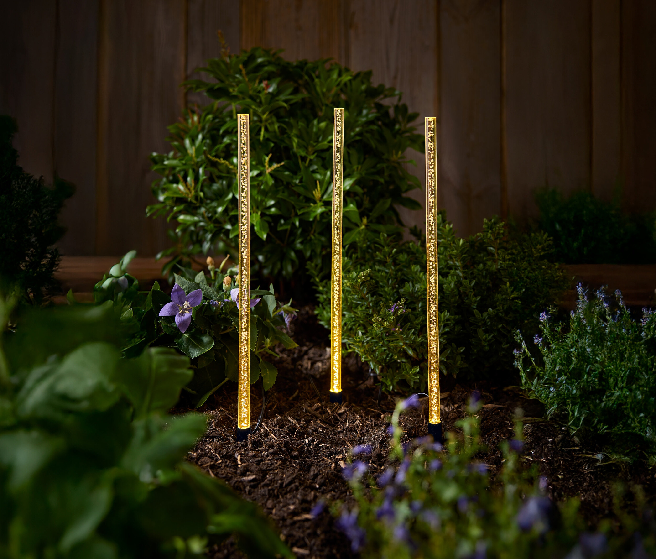 Tri zlaté solárne záhradné lampy stoja na záhone s kríkmi a kvetmi.