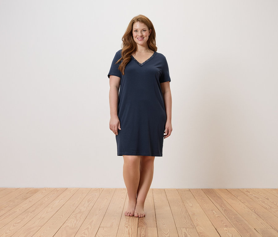 Žena stojí naboso na drevenej podlahe a má na sebe tmavomodrú nočnú košeľu.