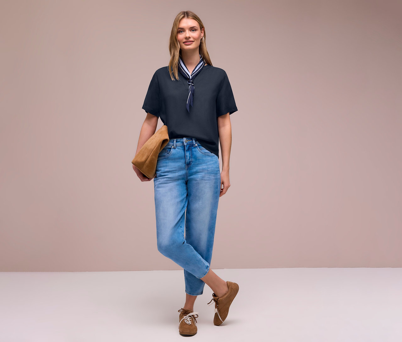 Žena v džínsoch a hnedých topánkach s taškou má na sebe tkanú blúzku Street One so šatkou.