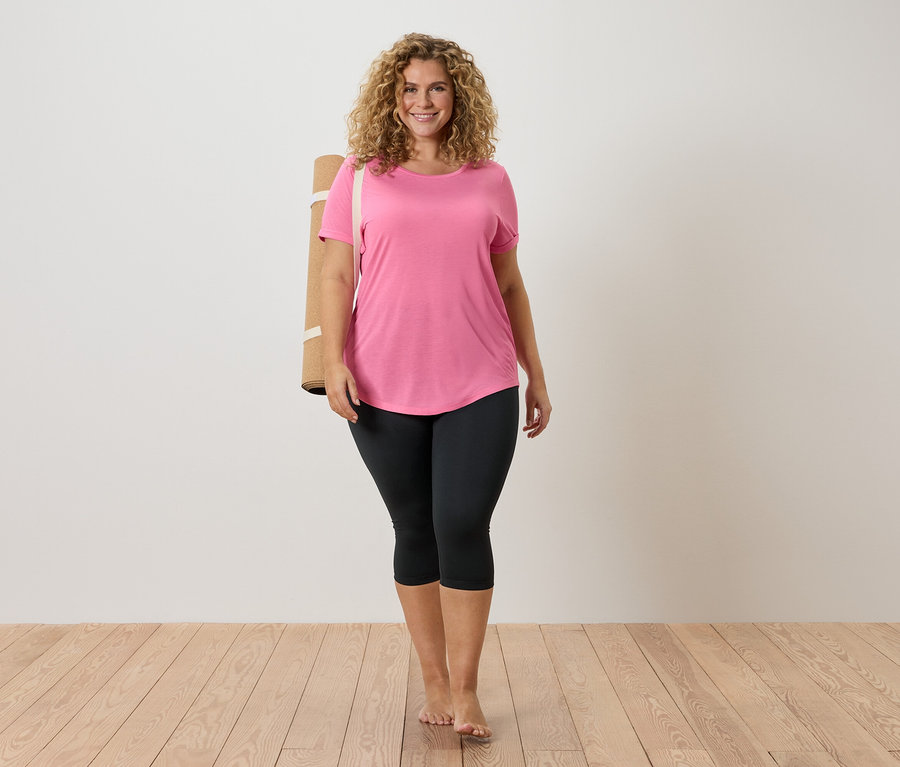 Žena s kučeravými vlasmi má na sebe ružové tričko a čierne legíny, s podložkou na jogu cez rameno, bosá na drevenej podlahe.