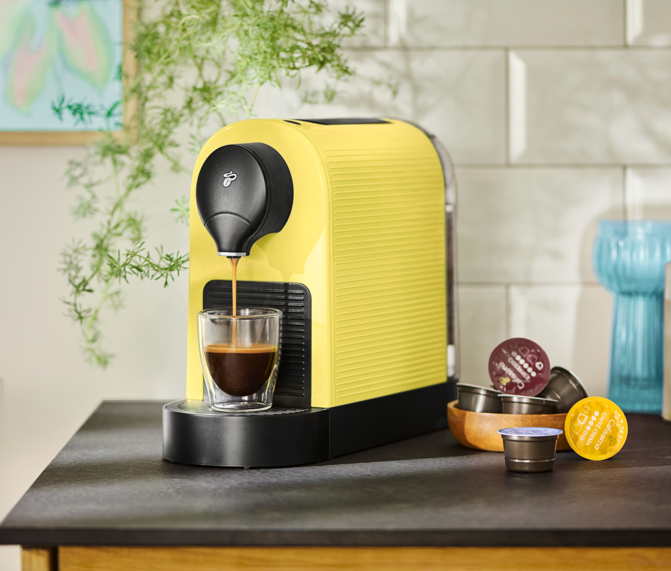 Žltý kávovar napĺňa šálku kávou, vedľa ležia kapsuly.