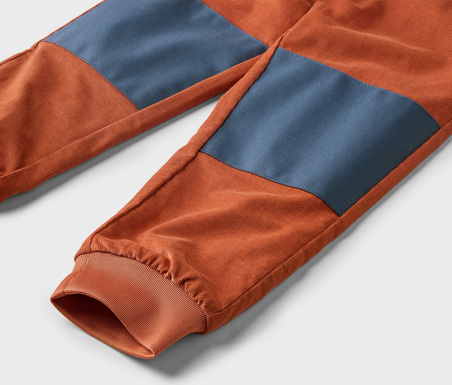 Detail oranžových menčestrových nohavíc s modrými záplatami na kolenách a elastickými manžetami na členkoch.
