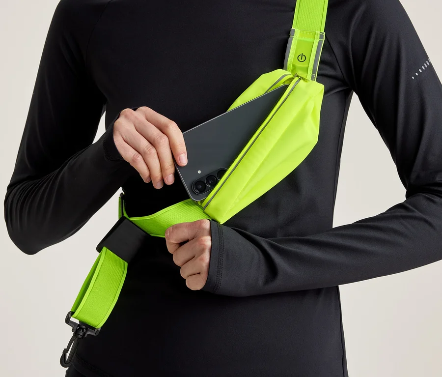 Osoba v čiernom tričku s dlhým rukávom vkladá čierny smartfón do neónovo žltej crossbody tašky s reflexným detailom.