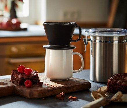 Čierny ručný filter na kávu veľkosti 101 stojí na bielom hrnčeku vedľa brownies s malinami na drevenej doske.