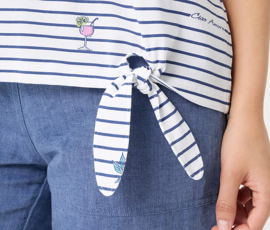 Detail osoby v bielo-modrom pruhovanom tričku Olsen, zaviazanom vpredu na uzol. Tričko má vyšívaný koktailový pohár, nápis 'Ciao Amore' a malý list na konci uzla, skombinované s modrými šortkami.