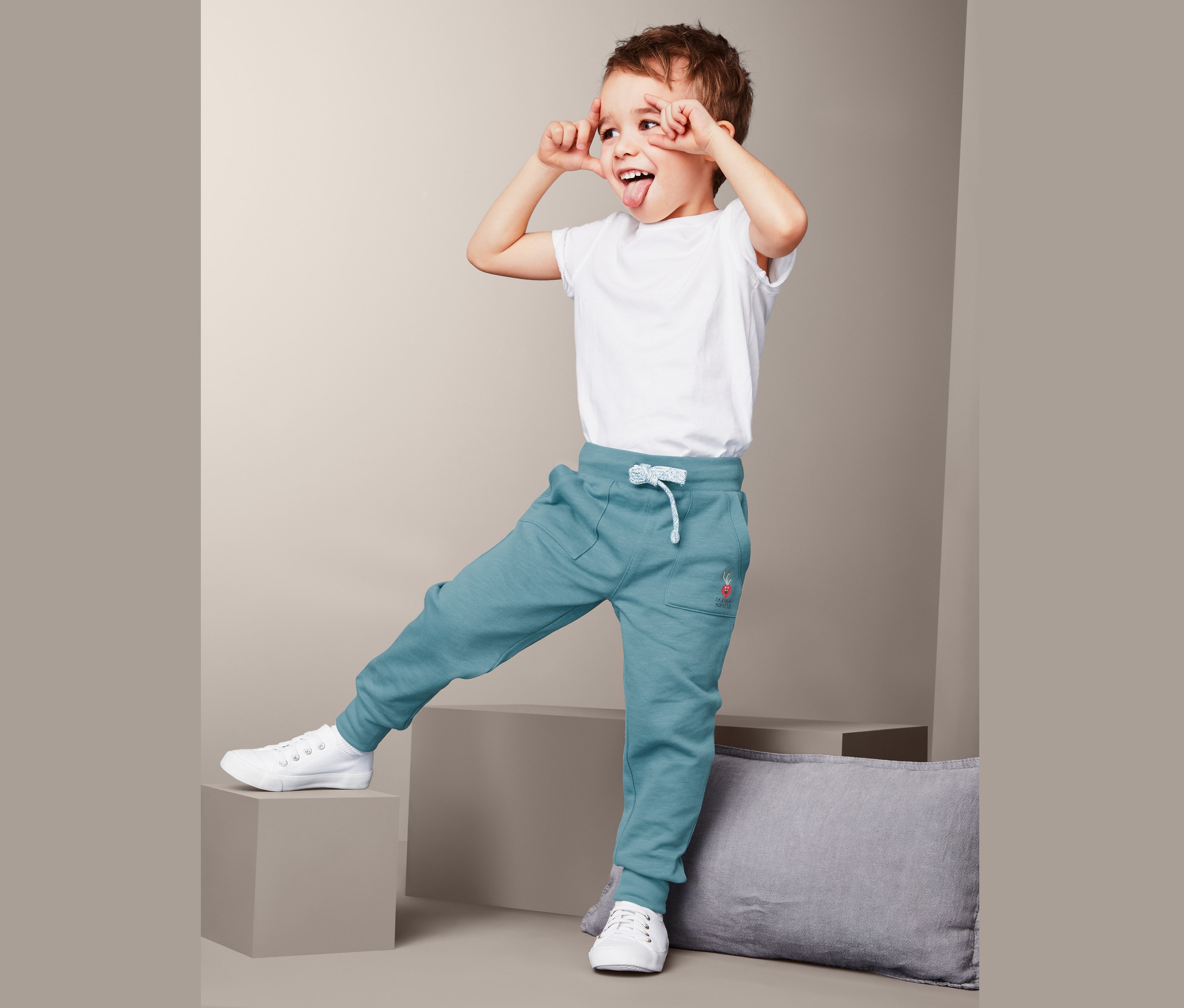 Chlapec v bielom tričku a teplákových nohaviciach stojí jednou nohou na sivom štvorci, vyplazuje jazyk a robí grimasy.