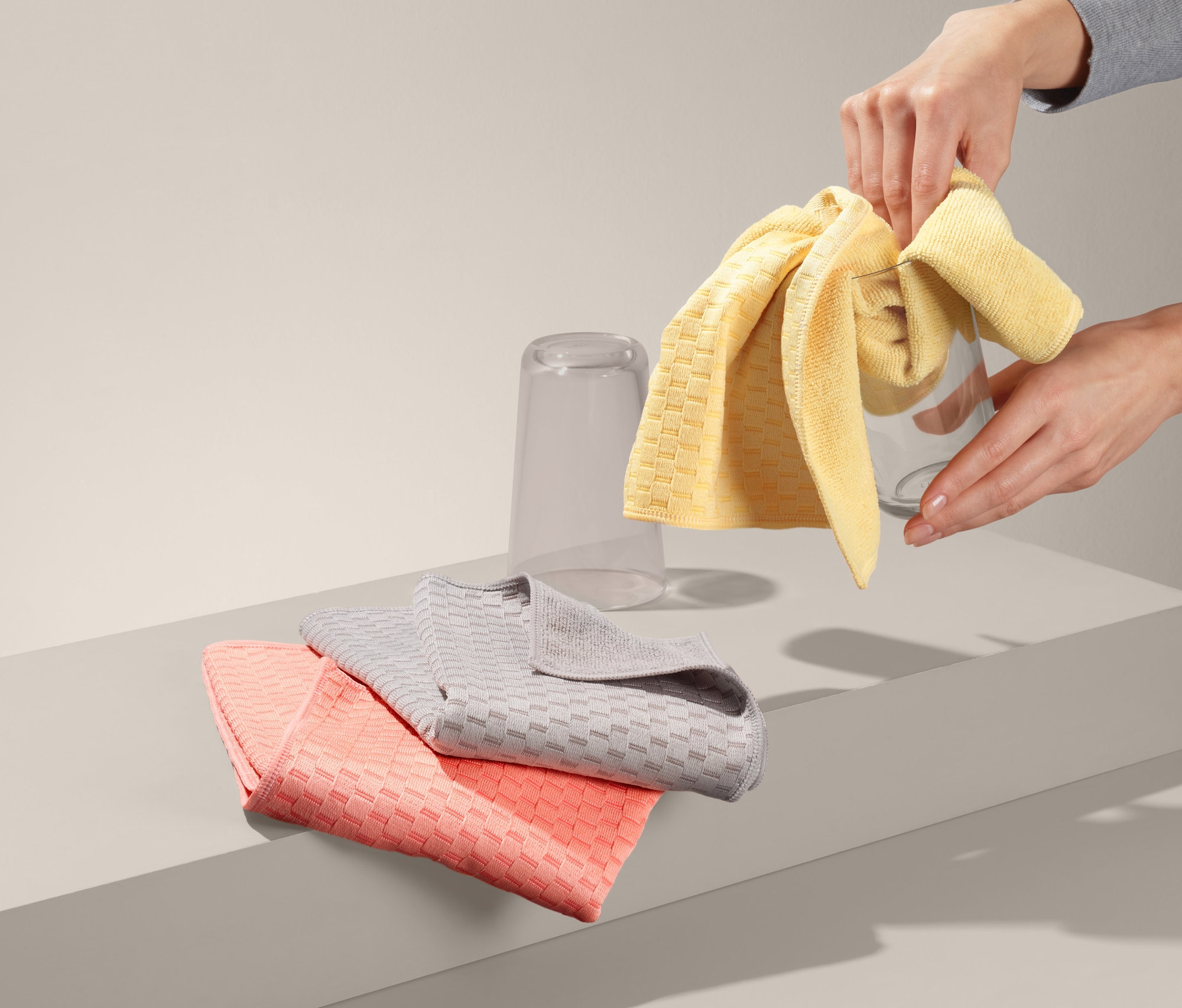 Ruky utierajú pohár žltou utierkou na riad. Na stole ležia zložené 3 špeciálne utierky na riad.