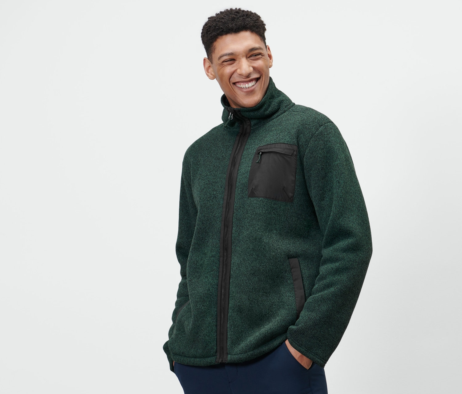 Muž má na sebe zelenú termobundu z pleteného flísu a tmavomodré nohavice.
