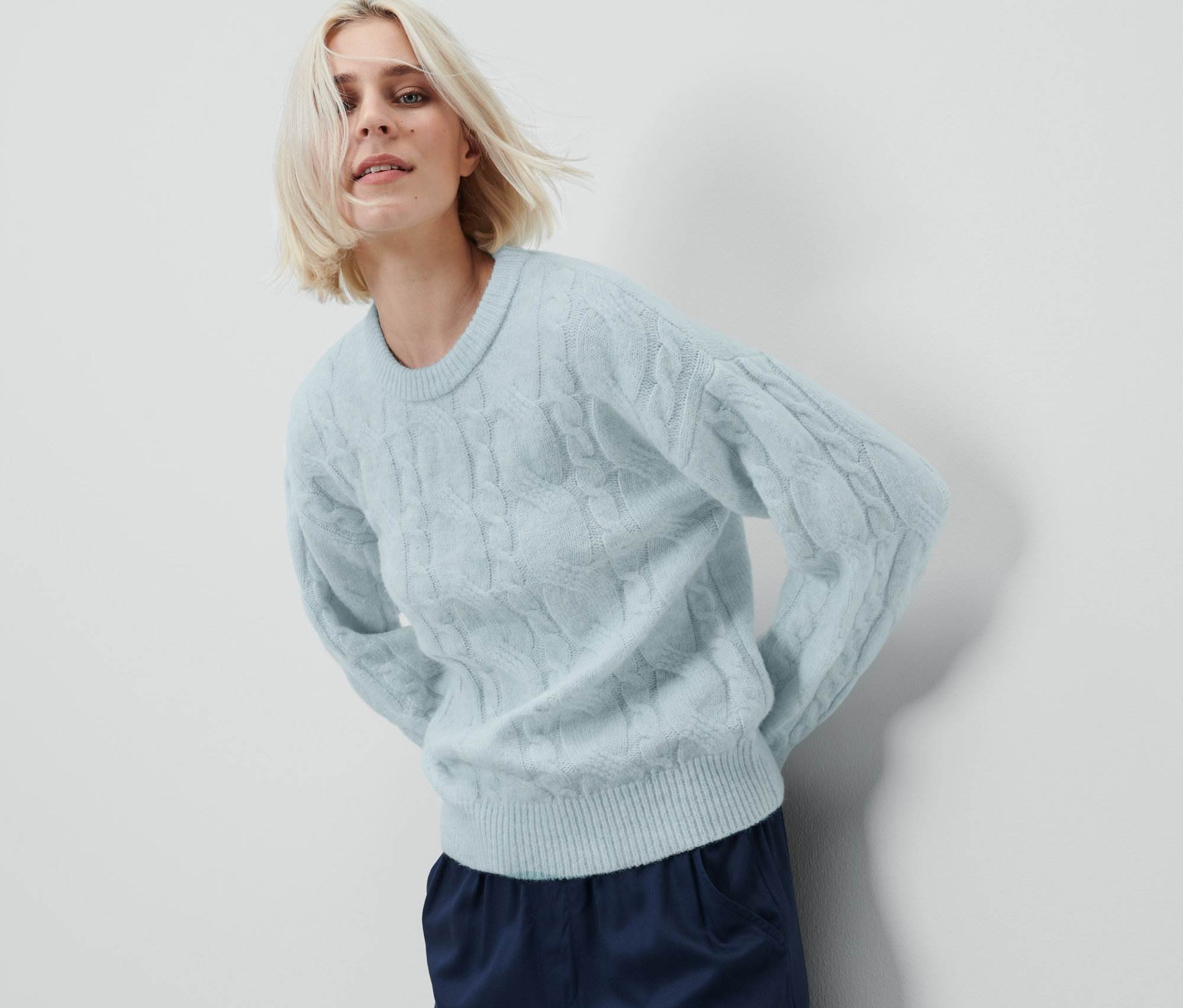 Žena pózuje na bielom pozadí a má na sebe svetlomodrý pletený pulóver.