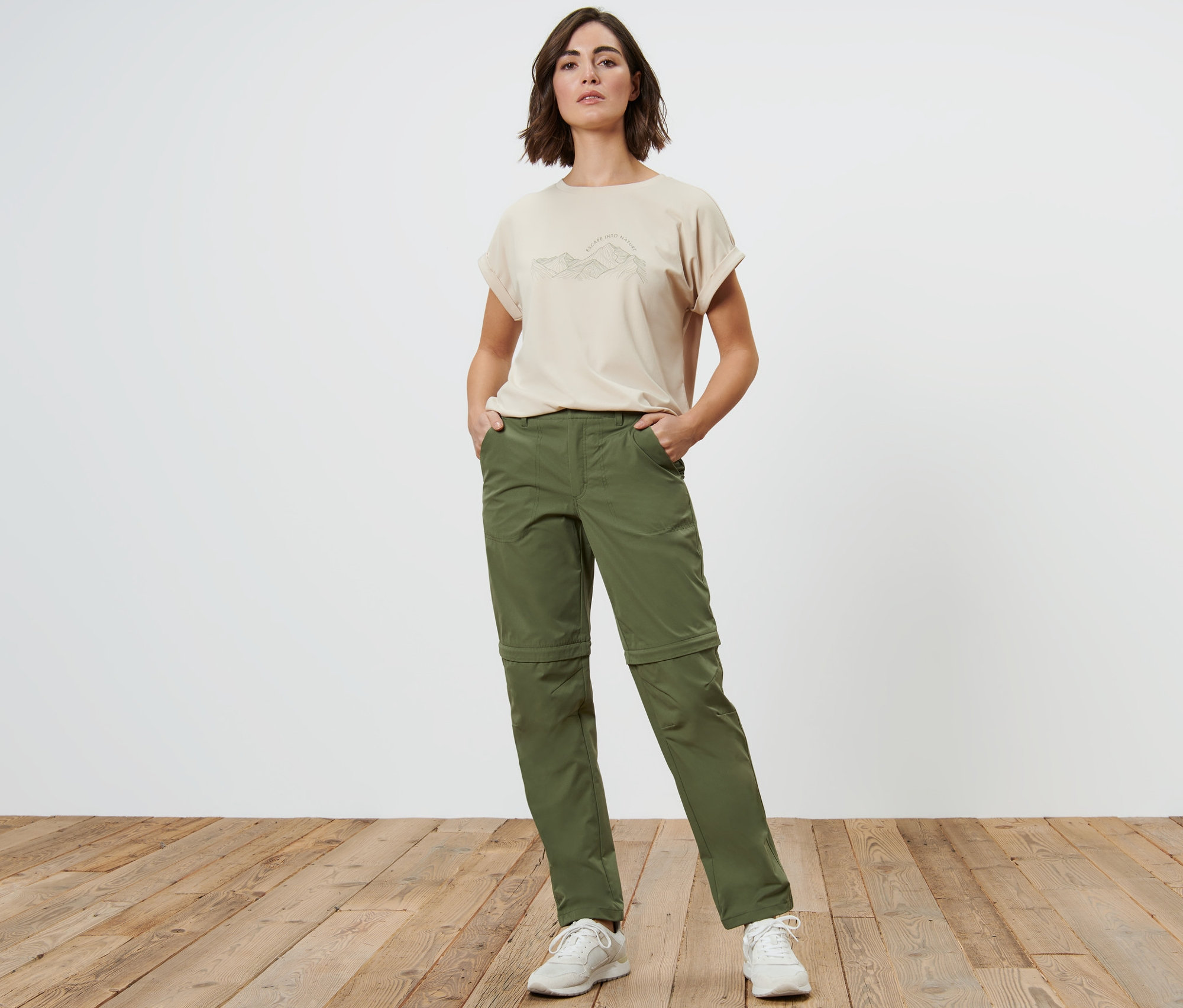 Žena stojí s rukami vo vreckách khaki funkčných nohavíc 2 v 1.