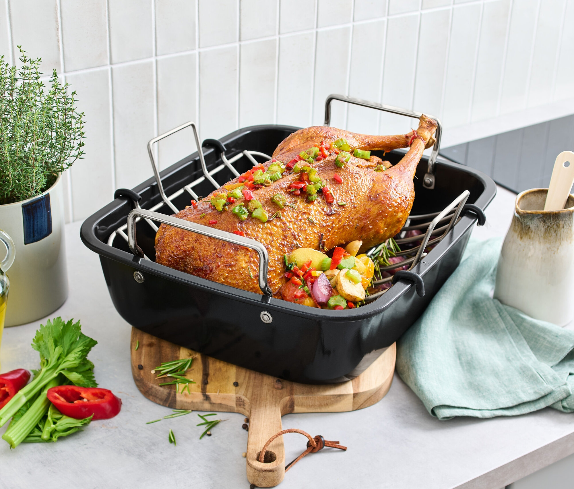 Pečená kačica v XL pekáči z ušľachtilej ocele s roštom, na stole so zeleninou a bylinkami.