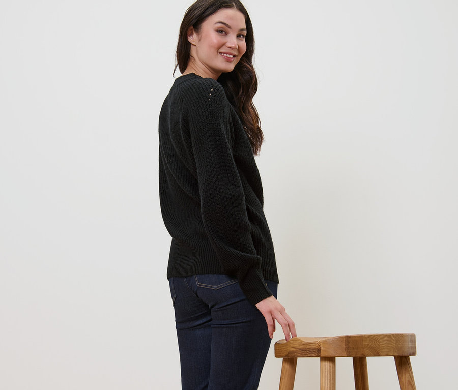 Žena sa usmieva cez plece a pózuje v čiernom pletenom pulóvri.