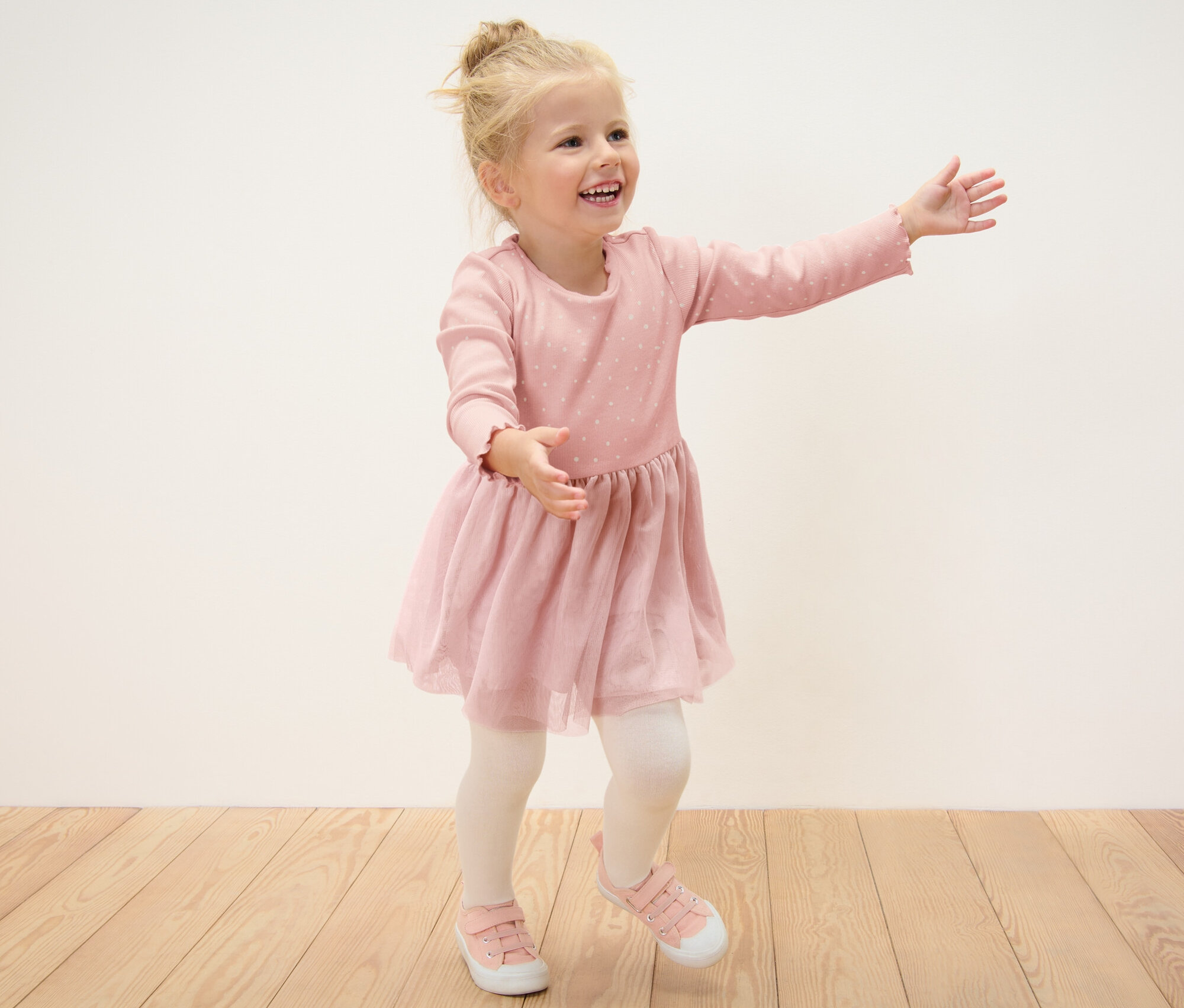 Usmievavé blond dievča v detských rebrovaných šatách pózuje na drevenej podlahe.