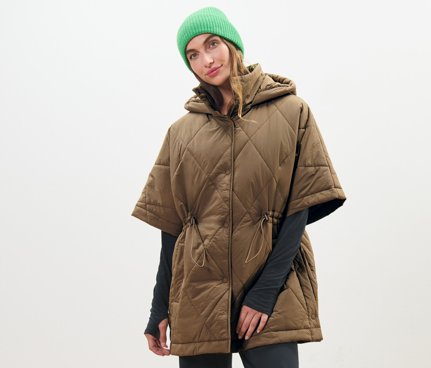 Žena nosí prešívané pončo s kapucňou a zelenú čiapku.