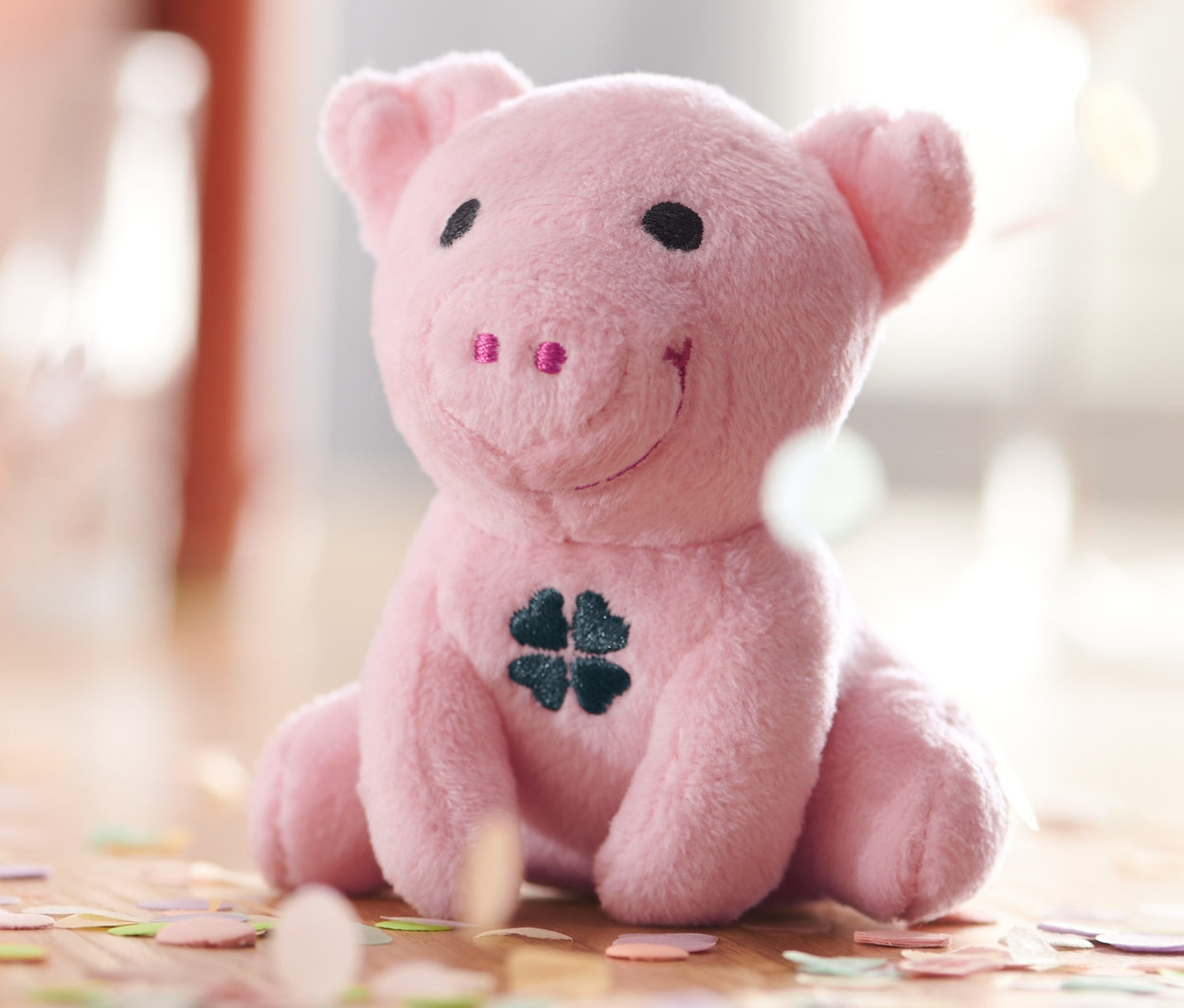 Sediace ružové prasiatko pre šťastie so zvukom a štvorlístkom na bruchu, obklopené konfetami.