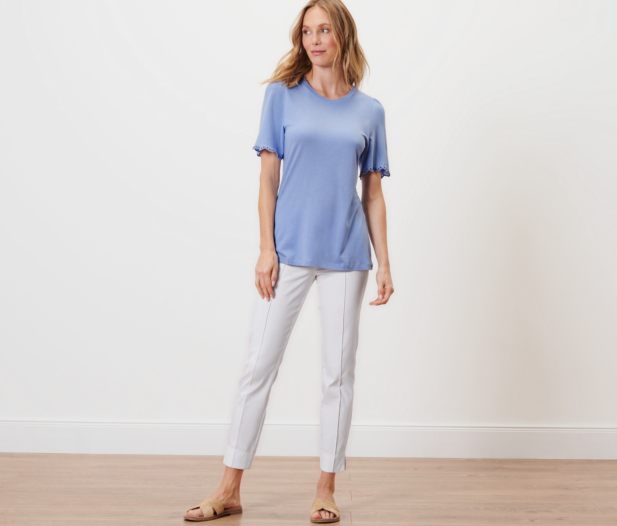 Žena stojí a má na sebe svetlomodré tričko s nariasením a biele nohavice.