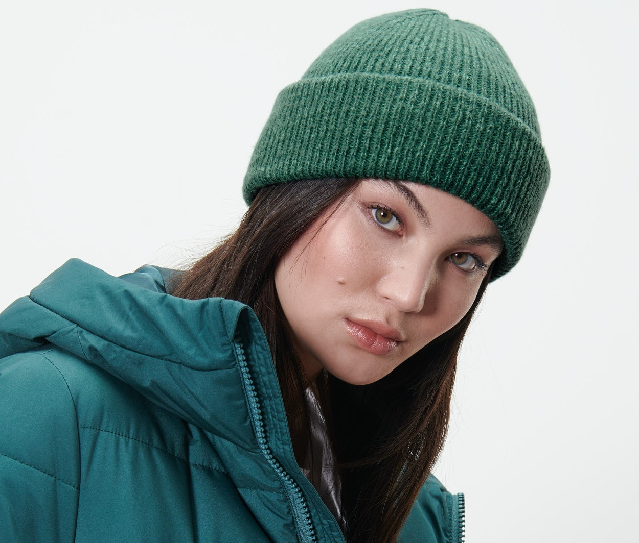 Žena má na sebe pletenú čiapku tmavozelenej farby a zelenú páperovú bundu.