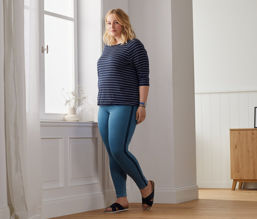 Blondína pózuje pri okne v tričku s netopierími rukávmi a v džegínsoch.