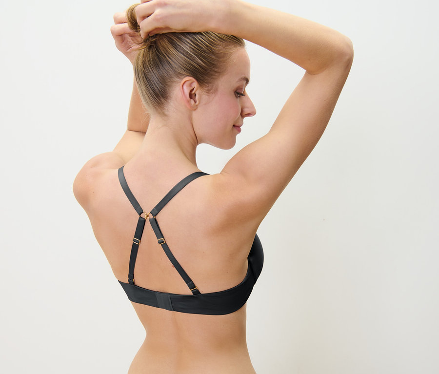 Blondínka si upravuje vlasy do drdola, na sebe má čiernu podprsenku s ozdobnými ramienkami.