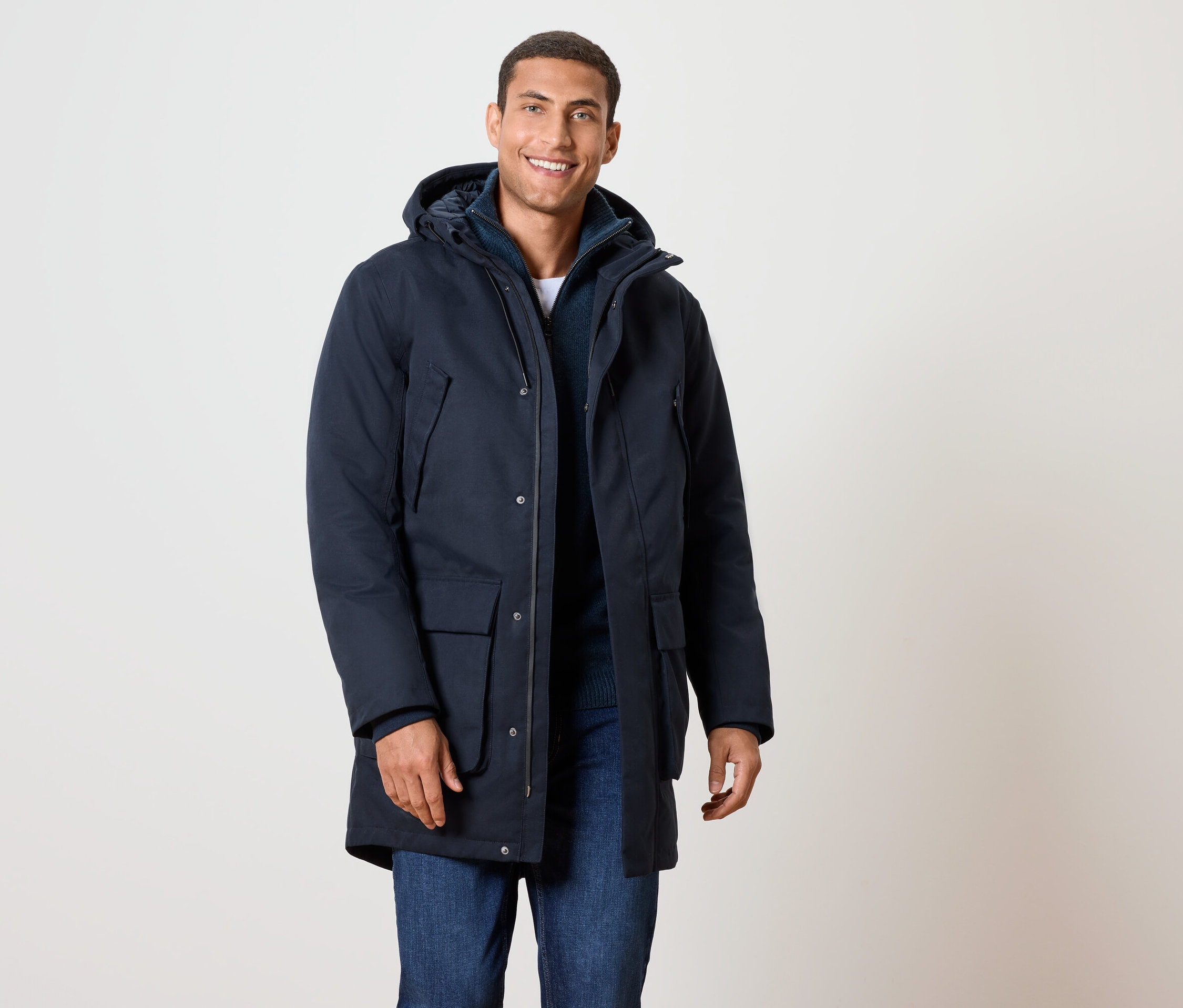 Usmievajúci sa muž má na sebe tmavomodrú zimnú bundu s kapucňou a džínsy.