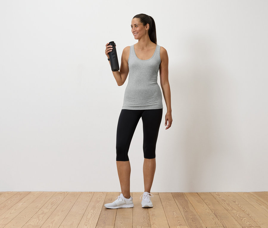 Žena drží fľašu s vodou, má na sebe sivé tielko, čierne legíny a biele tenisky.