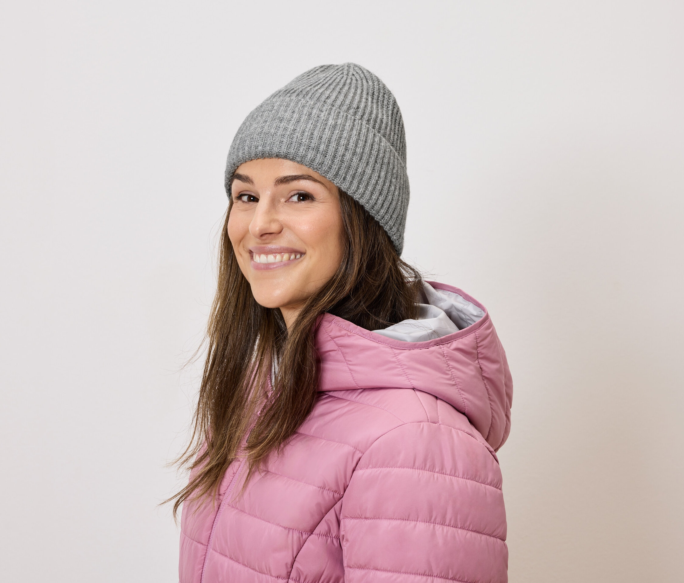 Žena s hnedými vlasmi sa usmieva a nosí sivú pletenú čiapku a ružovú páperovú bundu.
