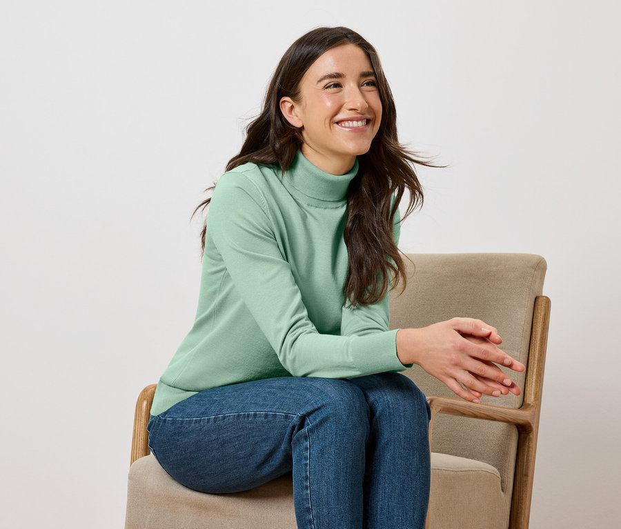 Usmievajúca sa žena sedí na béžovej stoličke, má na sebe zelený rolák a džínsy.