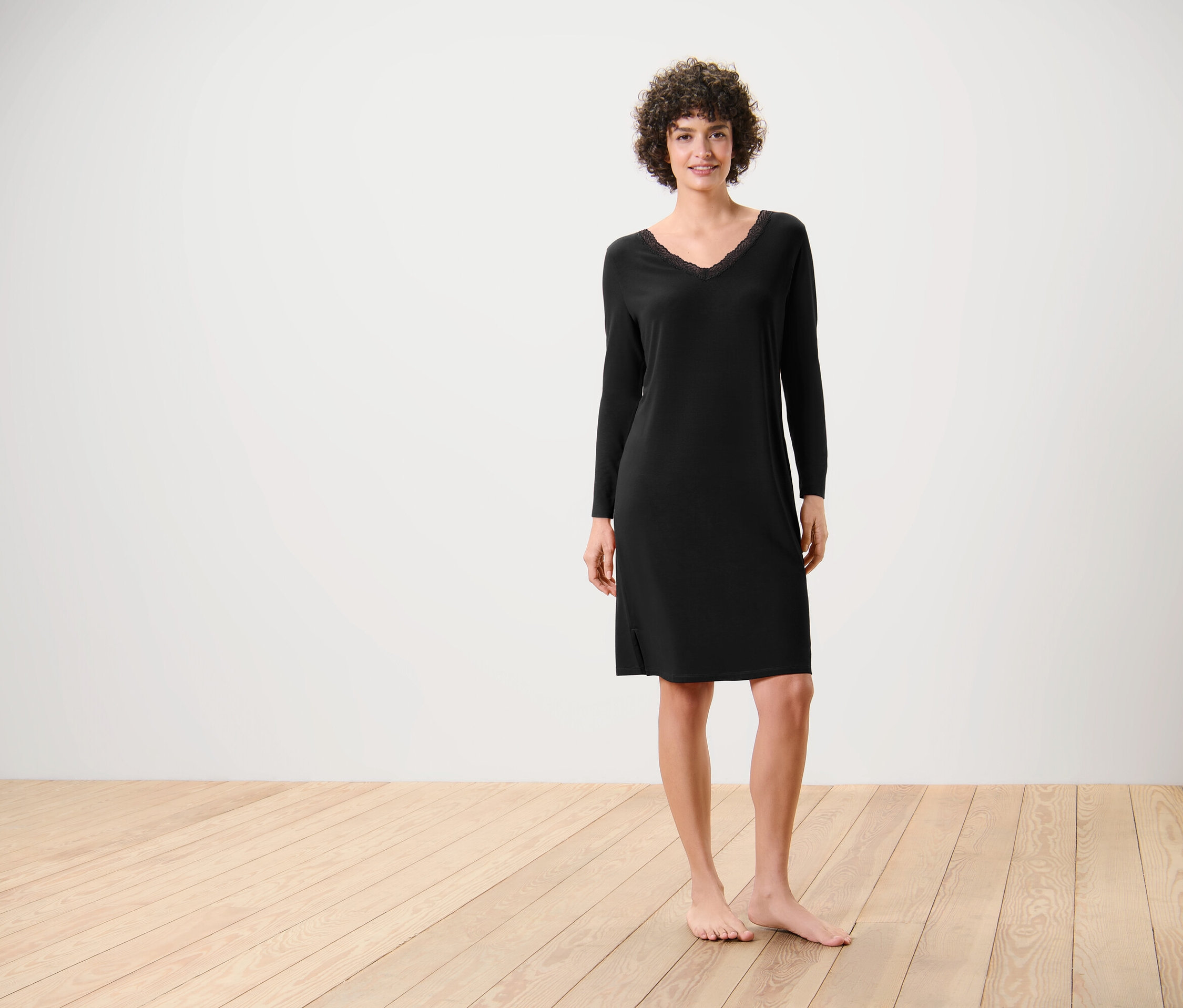 Žena stojí naboso na drevenej podlahe a má na sebe čierne šaty s dlhým rukávom.