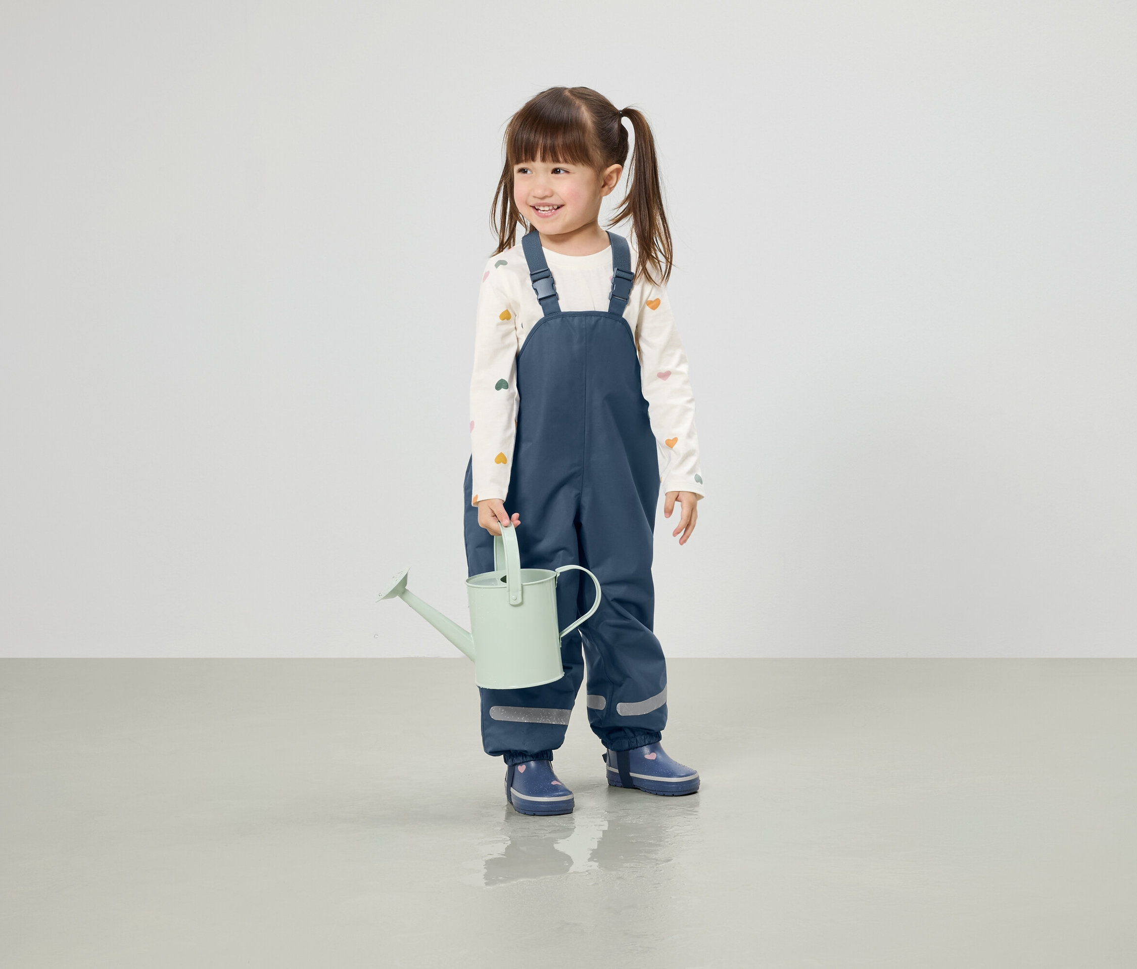 Usmievajúce sa dievča s vrkočmi drží svetlozelenú kanvicu, oblečené v modrých nohaviciach a gumákoch.