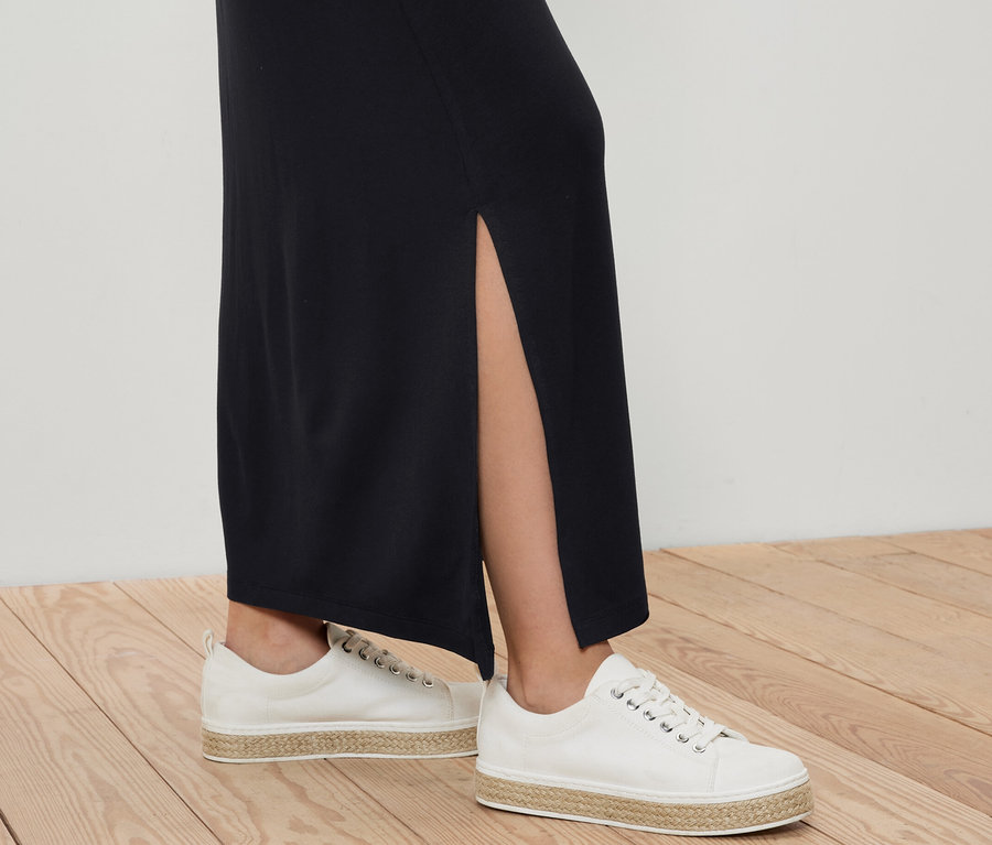 Nohy ženy v dlhej džersejovej sukni a bielych plátenných topánkach na drevenej podlahe.