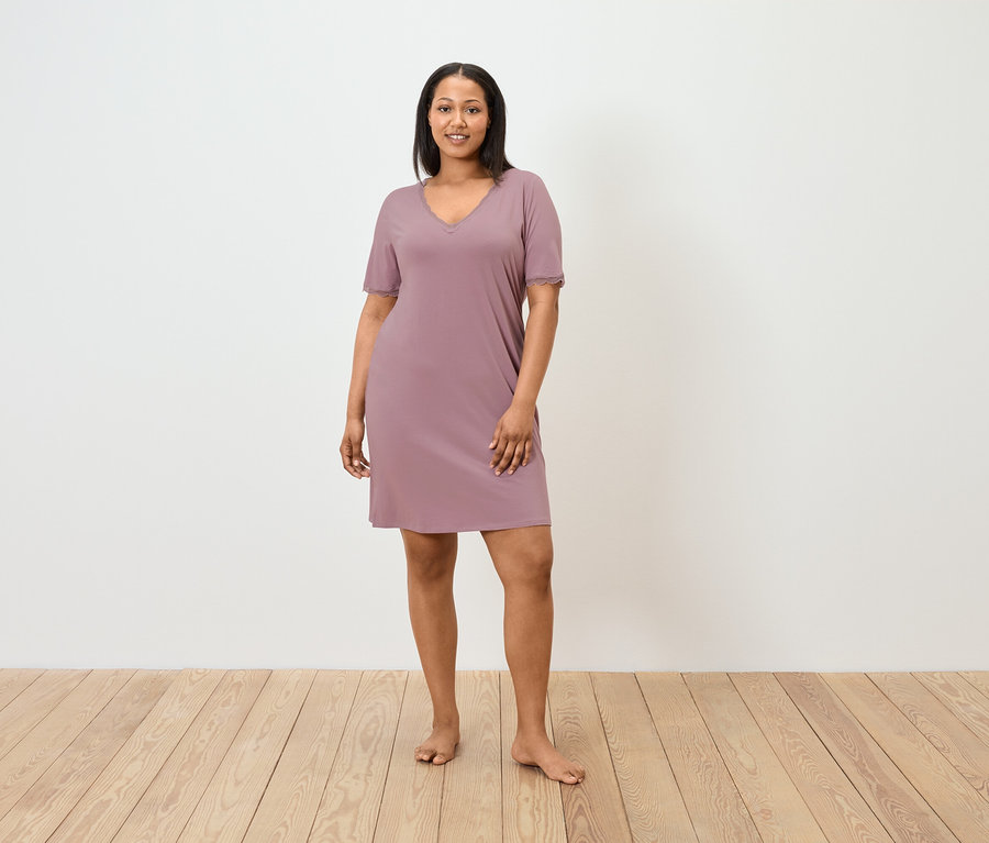 Žena stojí naboso a má na sebe krátku, fialovú nočnú košeľu.