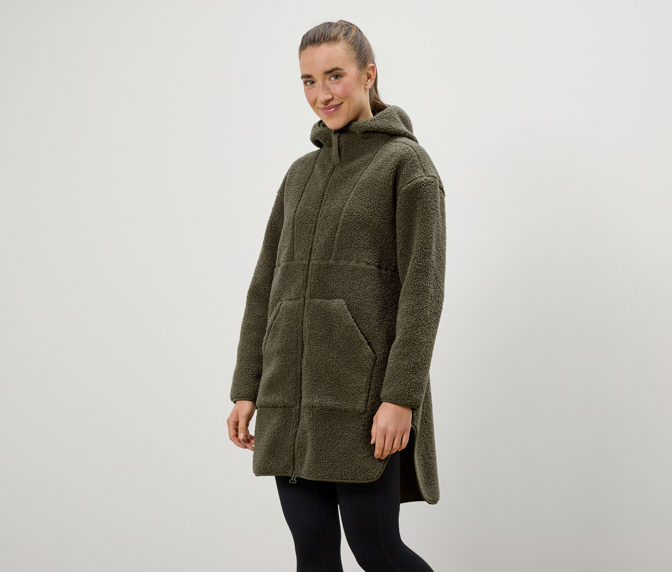 Žena má na sebe olivovo zelený kabát s kapucňou a čierne legíny.