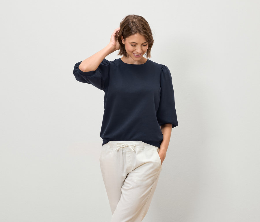 Žena pózuje v tmavomodrej blúzke s krátkymi rukávmi a svetlých nohaviciach.