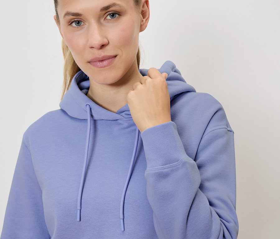 Portrét blondínky v lila mikine s kapucňou, ktorá si pridržiava kapucňu.