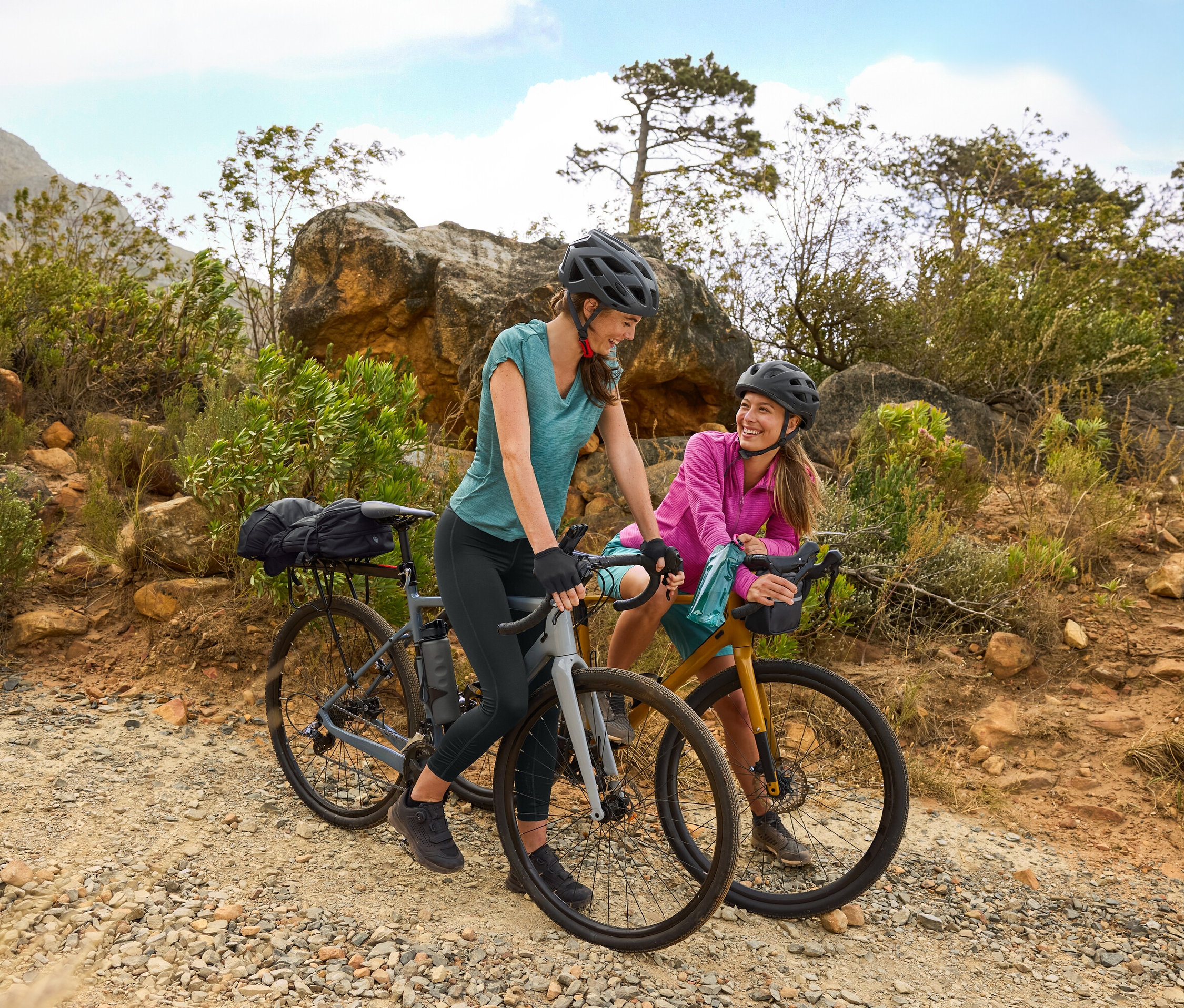 Dve ženy s prilbami stoja s bicyklami na štrkovej ceste v kopcovitom teréne. Na bicykli je pripevnená fľaša na pitie.