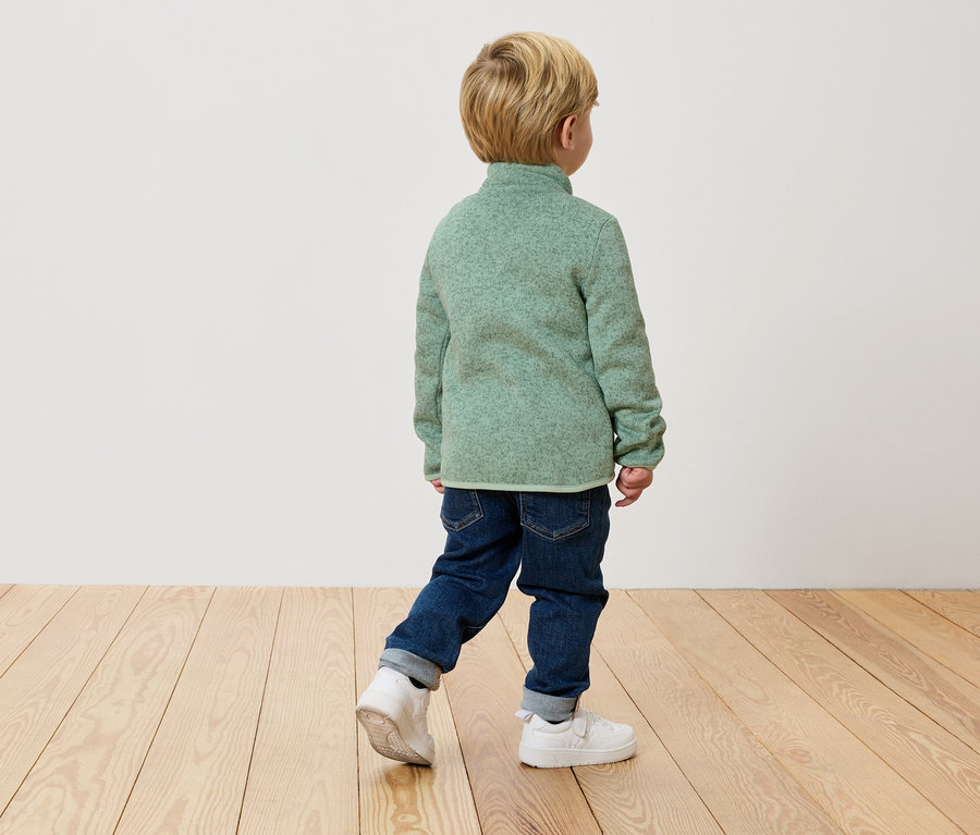 Dieťa kráča chrbtom k fotoaparátu v zelenom flísovom pulóvre, džínsoch a bielych teniskách na drevenej podlahe.