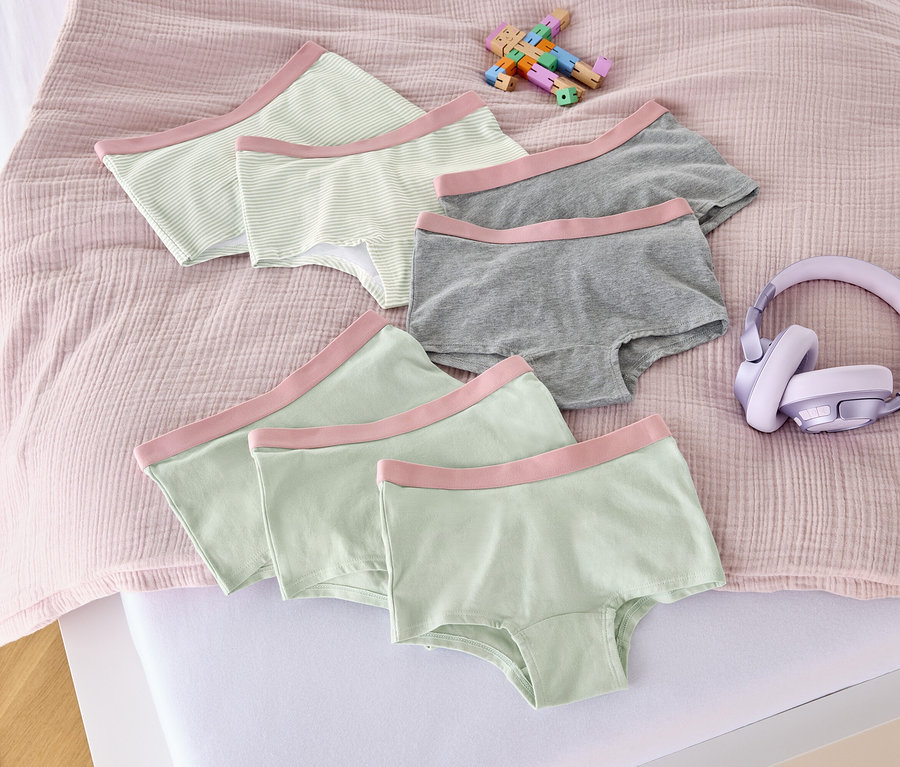 Na posteli leží sedem dievčenských nohavičiek v staroružovej farbe, hračka a slúchadlá.