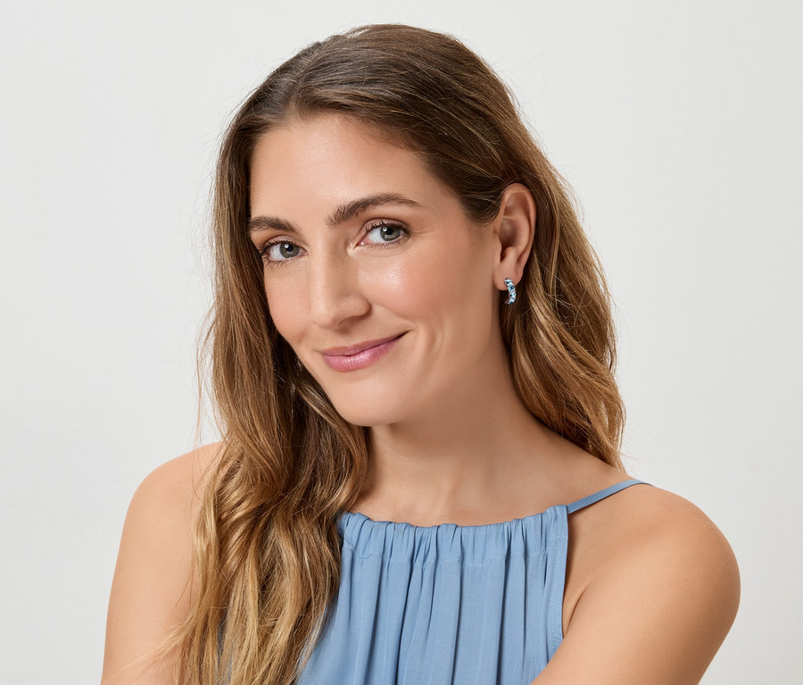 Žena s dlhými, vlnitými, hnedými vlasmi a v modrom tope sa usmieva do kamery a má na sebe modré náušnice.