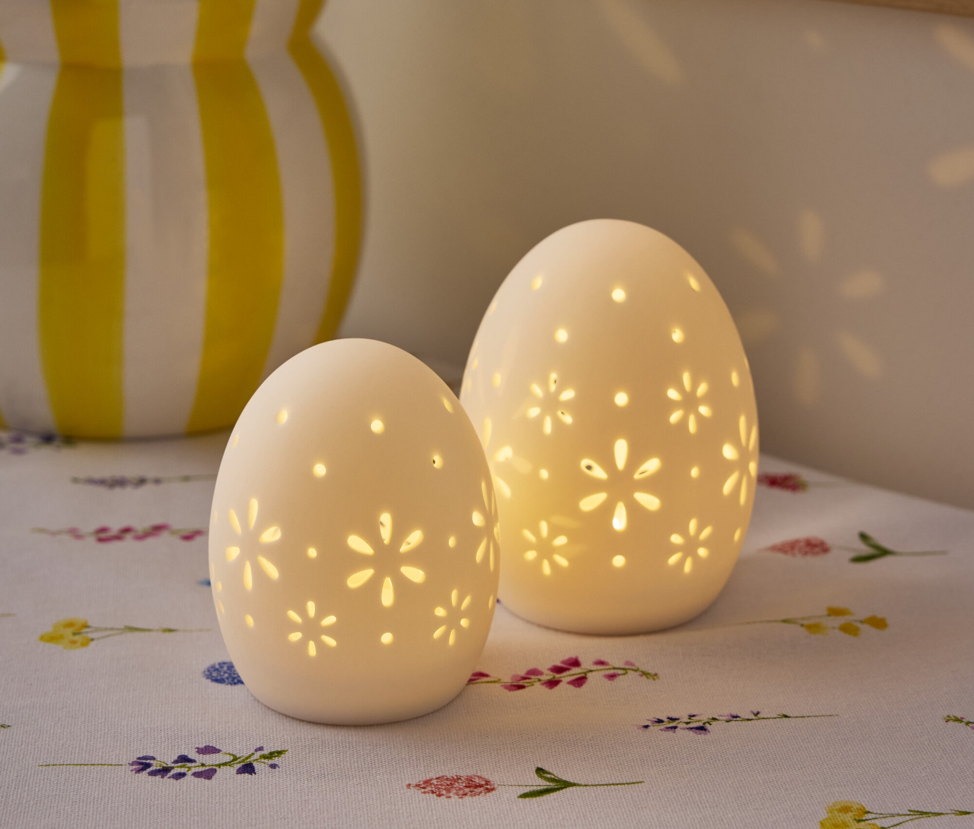 Dve biele lampy v tvare vajca s kvetinovým vzorom stoja na stole s vázou kvetov v pozadí.